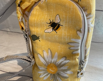 Op de afbeelding: Een gele canvas crossbody tas met een ritssluiting en een print van madeliefjes en bijen. De tas heeft een verstelbare grijze schouderband.