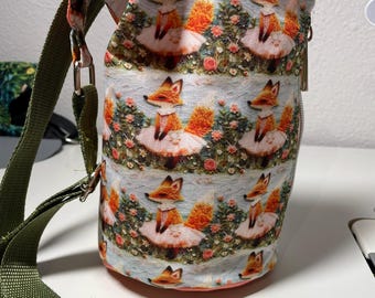 Op de afbeelding: Een cilindrische waterfleszak met een verstelbare groene riem. De tas is bedrukt met vossen in tutu's tussen bloemen. De binnenkant is koraalkleurig en de tas heeft een ritssluiting.