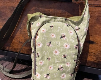 Op de afbeelding: Een groene canvas schoudertas met een patroon van madeliefjes en bijen. De tas heeft een ritssluiting en een verstelbare schouderband.