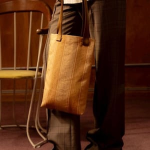 May include: A brown striped tote bag with a leather strap. The bag is being worn over the shoulder of a person wearing plaid trousers.