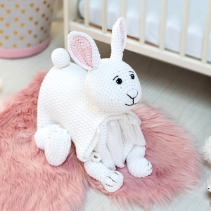 May include: White crocheted bunny rabbit toy with pink ears, sitting on a pink fluffy rug.