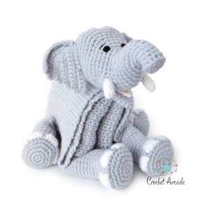 May include: A gray crocheted elephant stuffed animal with white accents. The elephant is sitting with its legs crossed and has a blanket draped over its back.