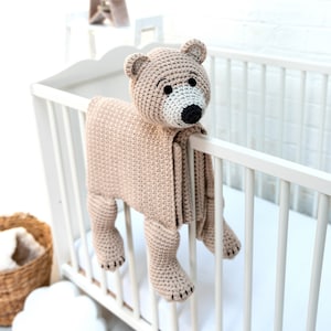 Pode incluir: Um brinquedo de urso de crochê marrom com nariz branco e olhos pretos pendurado em um berço branco. O urso está preso a um cobertor de crochê bege.