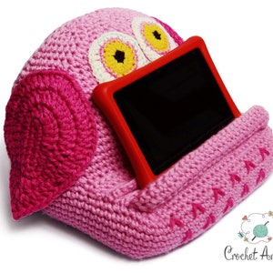 May include: Pink crocheted owl-shaped tablet stand with a red tablet case and a black screen. The stand has a crocheted owl face with yellow eyes and a pink beak.