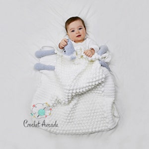 Pu&ograve; includere: Una copertina per neonati in crochet bianco con un agnello in crochet blu chiaro attaccato. La copertina ha un motivo testurizzato a punto bobble. Un bambino &egrave; sdraiato sulla copertina.