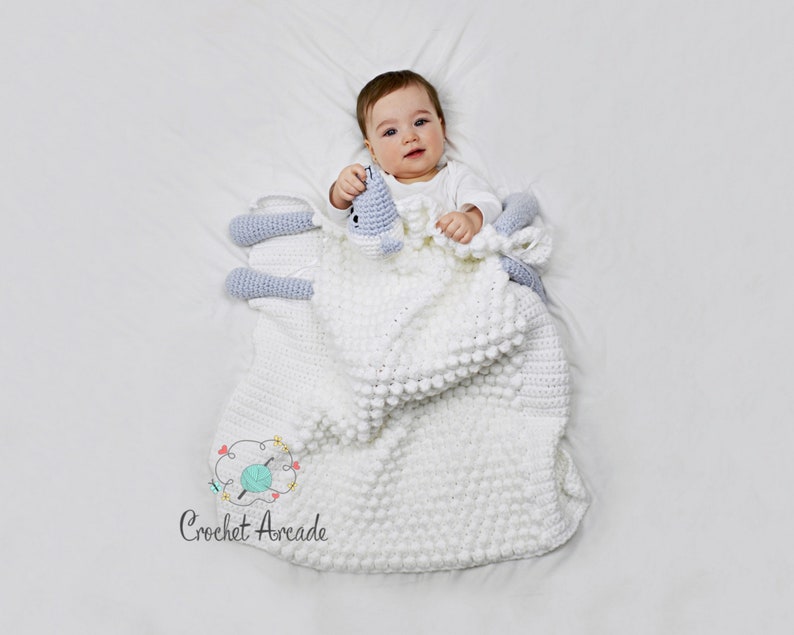 Pu&ograve; includere: Una copertina per neonati in crochet bianco con un agnello in crochet blu chiaro attaccato. La copertina ha un motivo testurizzato a punto bobble. Un bambino &egrave; sdraiato sulla copertina.