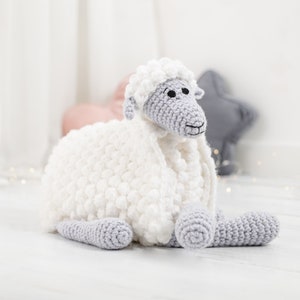 Pu&ograve; includere: Un giocattolo di pecora in crochet bianco con caratteristiche facciali e gambe grigie. La pecora &egrave; sdraiata su una superficie di legno bianca.