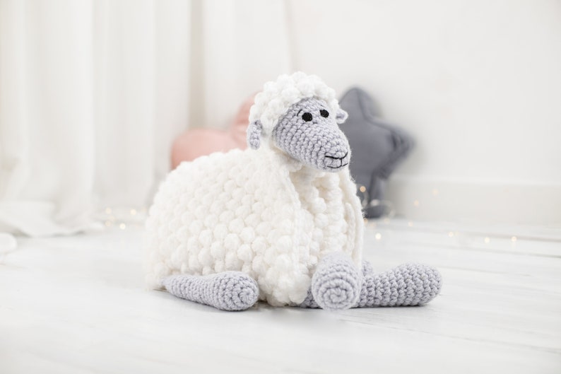 Pu&ograve; includere: Un giocattolo di pecora in crochet bianco con caratteristiche facciali e gambe grigie. La pecora &egrave; sdraiata su una superficie di legno bianca.