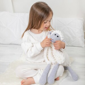 Pu&ograve; includere: Una bambina in pigiama bianco &egrave; seduta su un tappeto bianco e soffice e tiene un giocattolo di peluche a forma di pecora bianco e grigio lavorato a uncinetto.