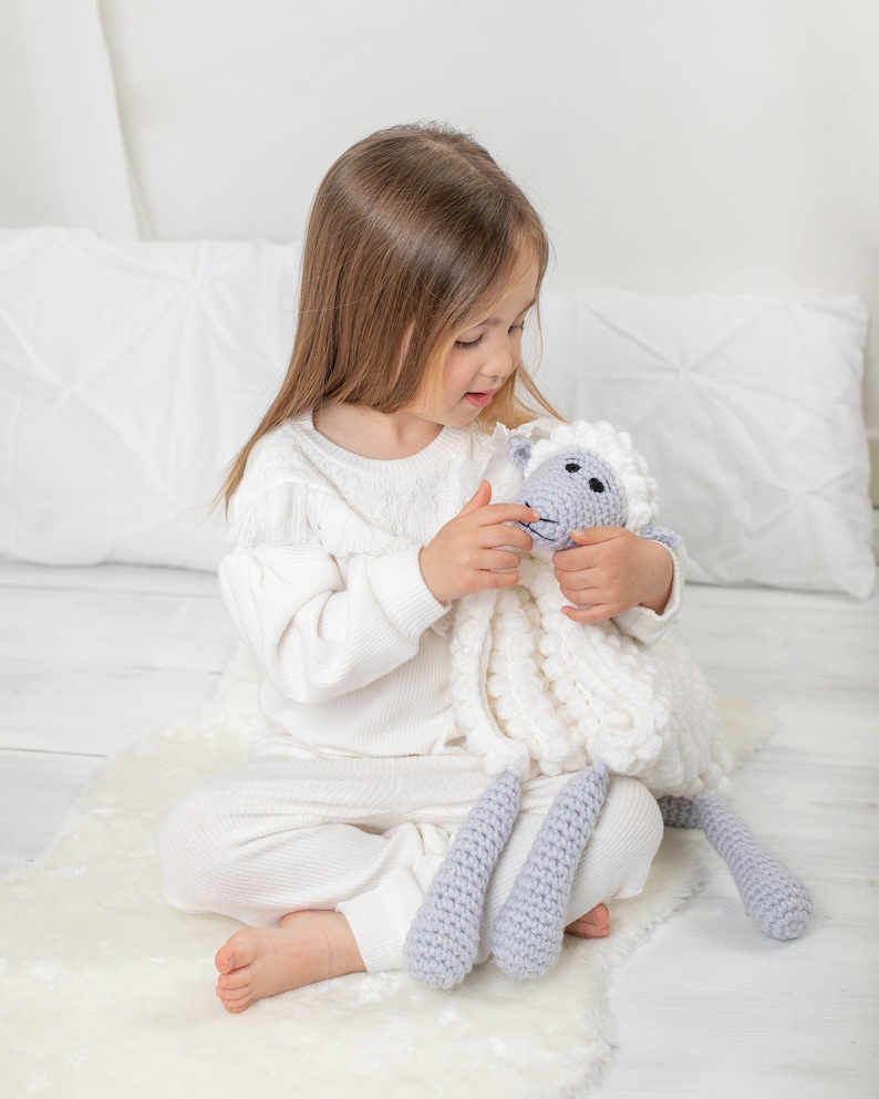 Pu&ograve; includere: Una bambina in pigiama bianco &egrave; seduta su un tappeto bianco e soffice e tiene un giocattolo di peluche a forma di pecora bianco e grigio lavorato a uncinetto.