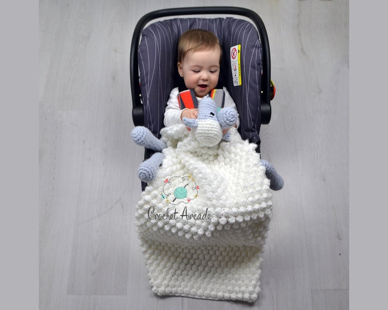 Pu&ograve; includere: Una copertina per neonati in crochet bianco con un animaletto di peluche in crochet grigio e bianco attaccato. La copertina &egrave; in un seggiolino auto con un bambino dentro.