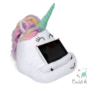 May include: White crocheted unicorn tablet holder with a rainbow mane and a green horn. The holder has a pocket for a tablet and a cute embroidered face.