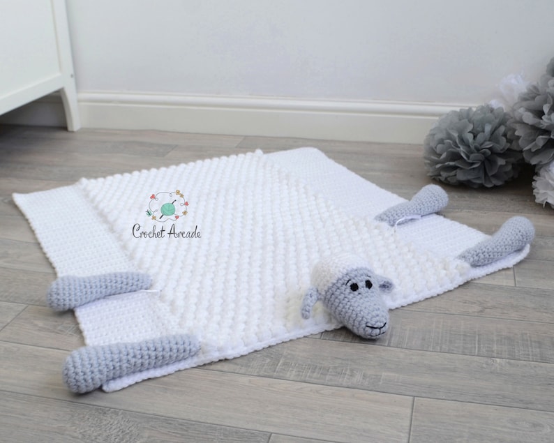 Pu&ograve; includere: Coperta per neonati in crochet bianco con una pecora in crochet grigio attaccata. La coperta ha una superficie testurizzata e irregolare. La pecora ha un viso grigio con occhi neri e un naso nero.