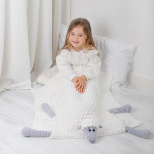 Pu&ograve; includere: Una coperta di lana bianca a forma di pecora con testa e gambe di pecora grigie. Una bambina &egrave; seduta sulla coperta, vestita con un vestito bianco.