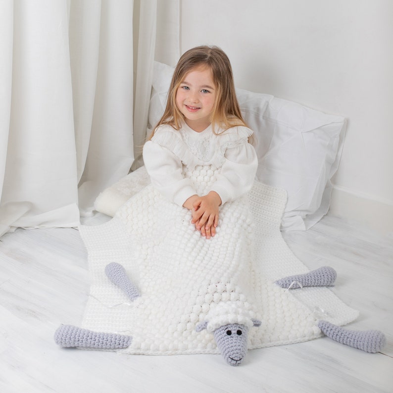 Pu&ograve; includere: Una coperta di lana bianca a forma di pecora con testa e gambe di pecora grigie. Una bambina &egrave; seduta sulla coperta, vestita con un vestito bianco.