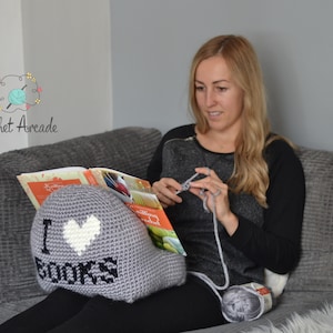 May include: A grey crocheted book holder with a white heart and the text "I love books" in black. The book holder is being used by a person sitting on a grey sofa.