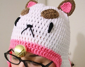 Pode incluir: Um chapéu de crochê branco e rosa em forma de gato, com orelhas marrons e um sino no topo. O chapéu tem um rosto de crochê branco com olhos pretos e um nariz.