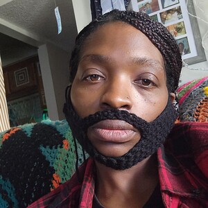 May include: A person wearing a red and black checked shirt with a black crocheted beard. The person has braided hair and is looking directly at the camera.