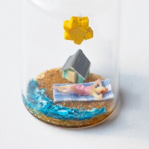 Op de afbeelding: Een miniatuur strandsc&egrave;ne in een glazen fles. Het bevat een klein figuur dat zonnebaadt op een blauw-wit handdoek, een klein huisje en een gele zon. De basis heeft zand en blauw water.