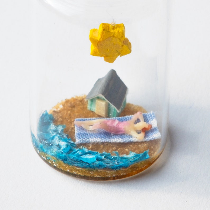 Op de afbeelding: Een miniatuur strandsc&egrave;ne in een glazen fles. Het bevat een klein figuur dat zonnebaadt op een blauw-wit handdoek, een klein huisje en een gele zon. De basis heeft zand en blauw water.