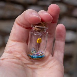 Op de afbeelding: Een miniatuur strandsc&egrave;ne in een heldere glazen fles met een kurk. De sc&egrave;ne bevat een persoon die zonnebaadt, een klein huis, een gele ster en blauw water. De fles wordt in een hand gehouden.