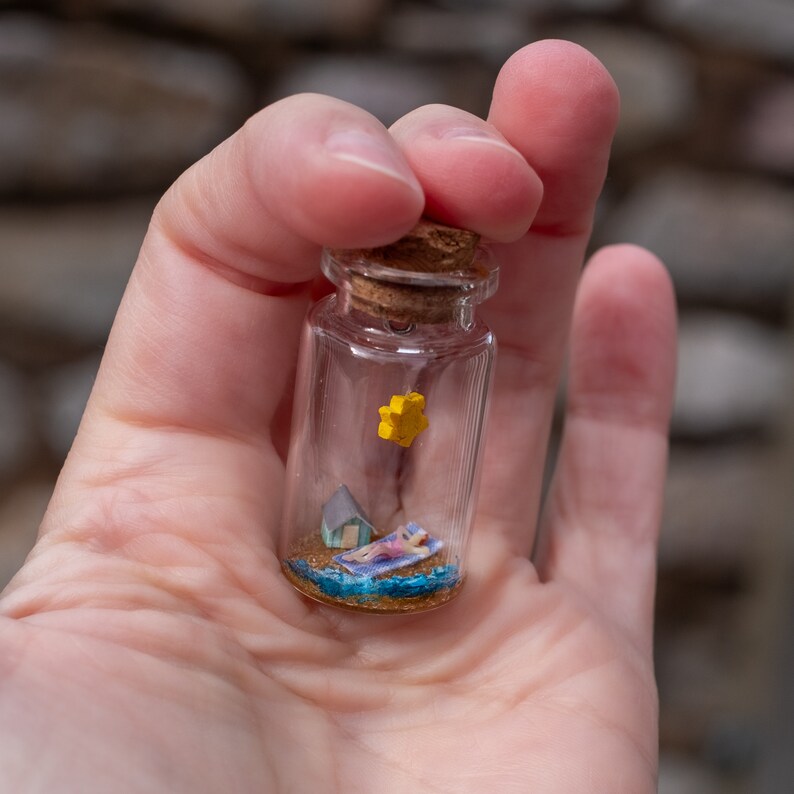Op de afbeelding: Een miniatuur strandsc&egrave;ne in een heldere glazen fles met een kurk. De sc&egrave;ne bevat een persoon die zonnebaadt, een klein huis, een gele ster en blauw water. De fles wordt in een hand gehouden.