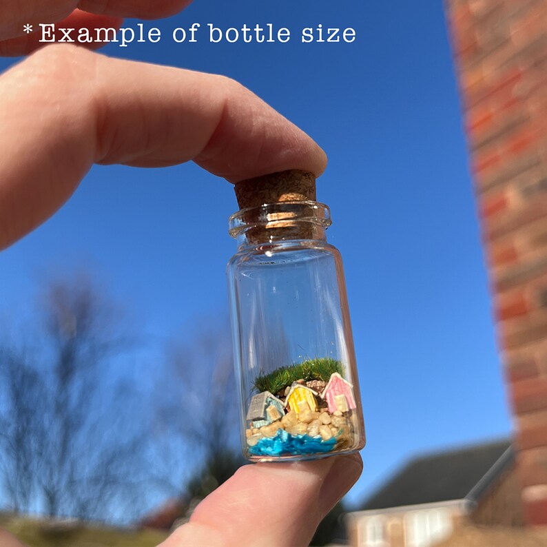 Op de afbeelding: Een kleine glazen fles met een kurk, met een miniatuur strandsc&egrave;ne met kleurrijke hutten, zand en blauw water. De tekst "Example of bottle size" is zichtbaar.