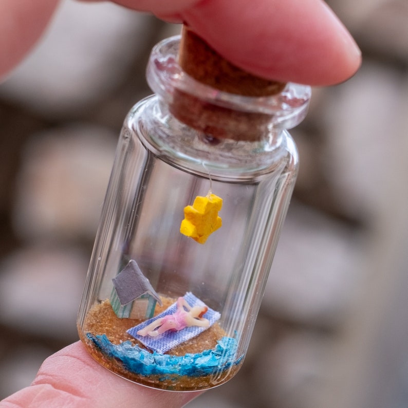 Op de afbeelding: Een miniatuur strandsc&egrave;ne in een heldere glazen fles. De sc&egrave;ne bevat een klein figuur dat zonnebaadt op een blauwe handdoek, een kleine strandhut en een gele ster die aan de kurk hangt. De fles wordt door een persoon vastgehouden.