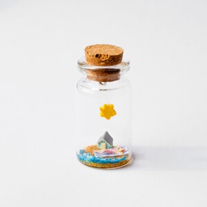 Op de afbeelding: Een miniatuur strandsc&egrave;ne in een heldere glazen fles met een kurk. De sc&egrave;ne bevat een klein huisje, een persoon die zonnebaadt en een gele ster. De fles is gevuld met zand en blauw water.