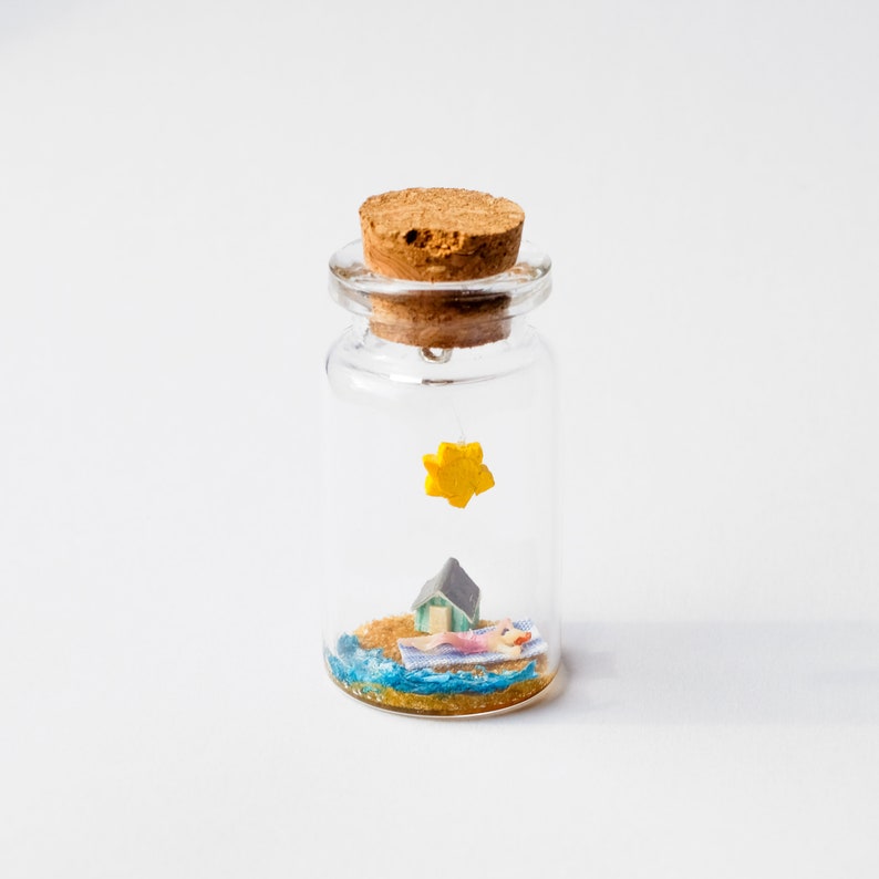 Op de afbeelding: Een miniatuur strandsc&egrave;ne in een heldere glazen fles met een kurk. De sc&egrave;ne bevat een klein huisje, een persoon die zonnebaadt en een gele ster. De fles is gevuld met zand en blauw water.