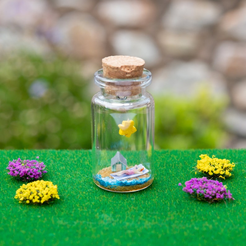 Op de afbeelding: Een heldere glazen fles met een kurk, met een miniatuur strandsc&egrave;ne. Binnenin zijn een klein huis, een persoon die zonnebaadt en een gele ster zichtbaar. De fles is omgeven door kleine paarse en gele bloemaccenten op een groen oppervlak.