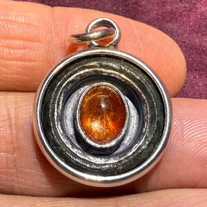 May include: A silver pendant with a circular design. The pendant features concentric rings surrounding an oval, orange-brown gemstone. The gemstone has a shimmering, reflective quality. The pendant is designed to be worn as jewelry.