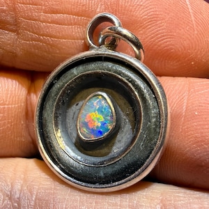 May include: A silver pendant with a circular, layered design. The center features a vibrant, oval-shaped opal with a spectrum of colors. The pendant has a loop for a chain and is held by a hand.