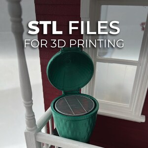 May include: A green 3D-printed miniature grill with an open lid, revealing a grill grate. The text "STL FILES FOR 3D PRINTING" is displayed above the grill. The grill is set against a red wall and a white window frame.