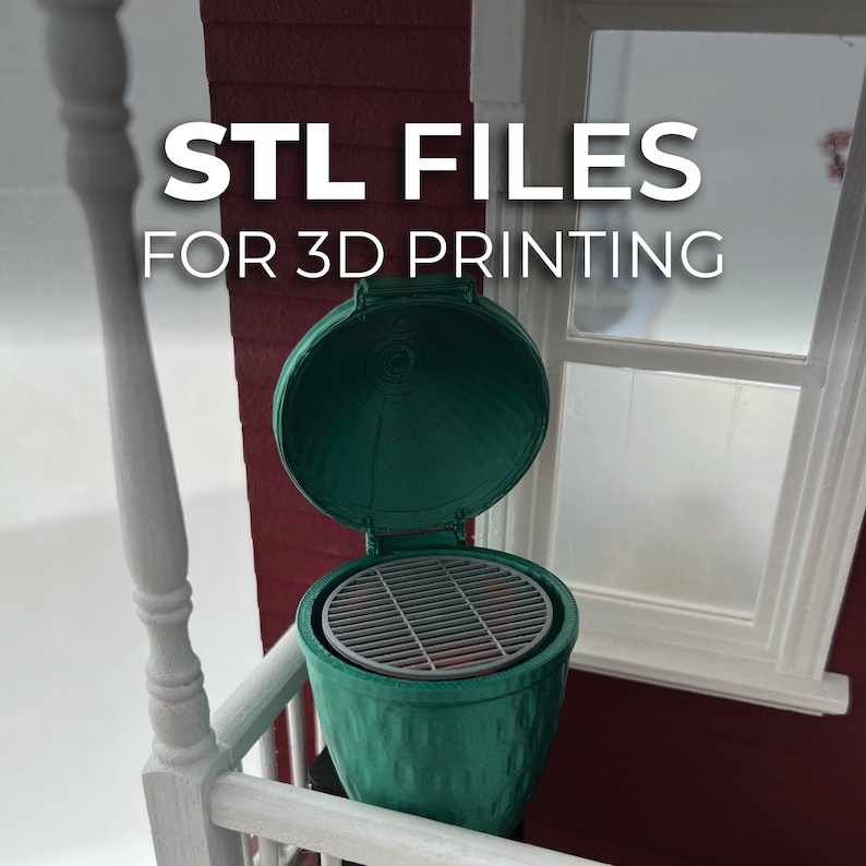 May include: A green 3D-printed miniature grill with an open lid, revealing a grill grate. The text "STL FILES FOR 3D PRINTING" is displayed above the grill. The grill is set against a red wall and a white window frame.