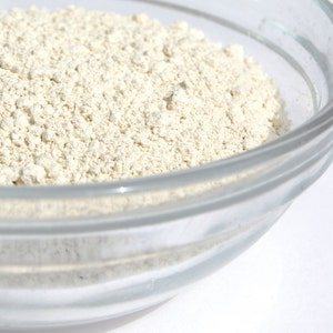 May include: A clear glass bowl filled with a light beige, finely ground powder. The powder has a slightly textured appearance, with small granules visible. The bowl is set against a bright white background, highlighting the contents.