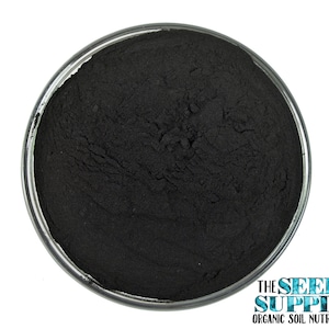 May include: A close-up of a small glass dish filled with a fine black powder. The powder is smooth and uniform in texture. The dish is sitting on a white background.