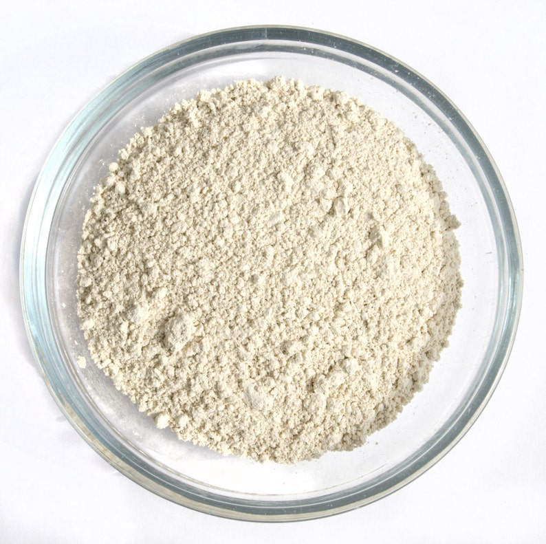 May include: A clear glass bowl filled with a light beige, finely ground powder. The powder has a slightly textured appearance and fills the bowl to the top. The bowl is set against a bright white background.