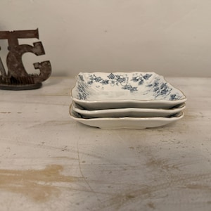 May include: A stack of three white, square-shaped ceramic dishes with a blue floral pattern and gold trim. The dishes are stacked on top of each other. In the background is a metal sculpture of the letters "TG".