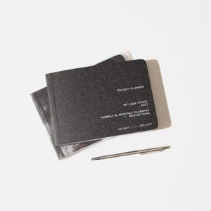 May include: A black pocket planner with a textured cover and white text that reads POCKET PLANNER BY CASE STUDY 2027 WEEKLY & MONTHLY PLANNING + REFLECTIONS JAN 2027 - DEC 2027, accompanied by a slim silver metal pen positioned below the planner.