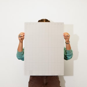 May include: A large, rectangular, off-white cutting mat with a grid pattern and measurement markings. The mat is held up by a person wearing a teal sweatshirt and brown pants. The mat is designed for crafting or sewing projects.