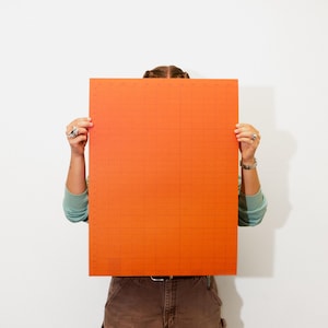 May include: A large, orange rectangular grid chart held by a person. The chart has a grid pattern with horizontal and vertical lines. The person is wearing a teal long-sleeved shirt and brown pants.