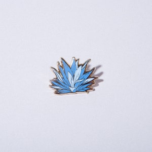 May include: A gold-colored pin with a blue and white agave plant design.