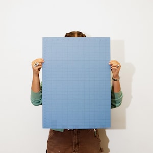 May include: A light blue cutting mat with a grid pattern and measurement markings. The mat is held up in front of a person, obscuring their face. The mat is rectangular and appears to be made of a durable material.