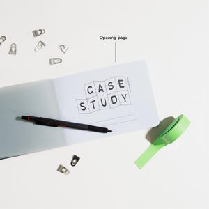 Peut inclure: Un cahier ouvert avec les mots "CASE STUDY" imprim&eacute;s sur la page. Un stylo noir, du ruban adh&eacute;sif vert et plusieurs trombones argent&eacute;s sont &eacute;galement sur la surface blanche. Le cahier est ouvert &agrave; la page d'ouverture.