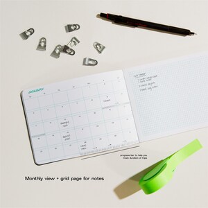 Peut inclure: Un cahier ouvert avec un calendrier mensuel et une page quadrill&eacute;e pour les notes. Un stylo noir, des trombones et un ruban vert sont sur la surface blanche. Le texte indique 'Vue mensuelle + page quadrill&eacute;e pour les notes'.