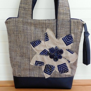 May include: A brown and blue tote bag with a large flower applique made of beige fabric and blue trim. The flower has a cluster of blue buttons in the center.