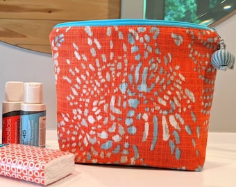 May include: A bright orange and blue patterned zippered pouch with a blue tassel. The pouch is sitting on a white surface with four small bottles and a package of tissues.
