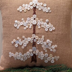 May include: A burlap pillow with a white button Christmas tree design. The tree is made of white buttons of various sizes and shapes.
