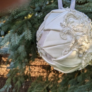 May include: A white fabric Christmas ornament with a beaded and lace design. The ornament is hanging from a tree branch.